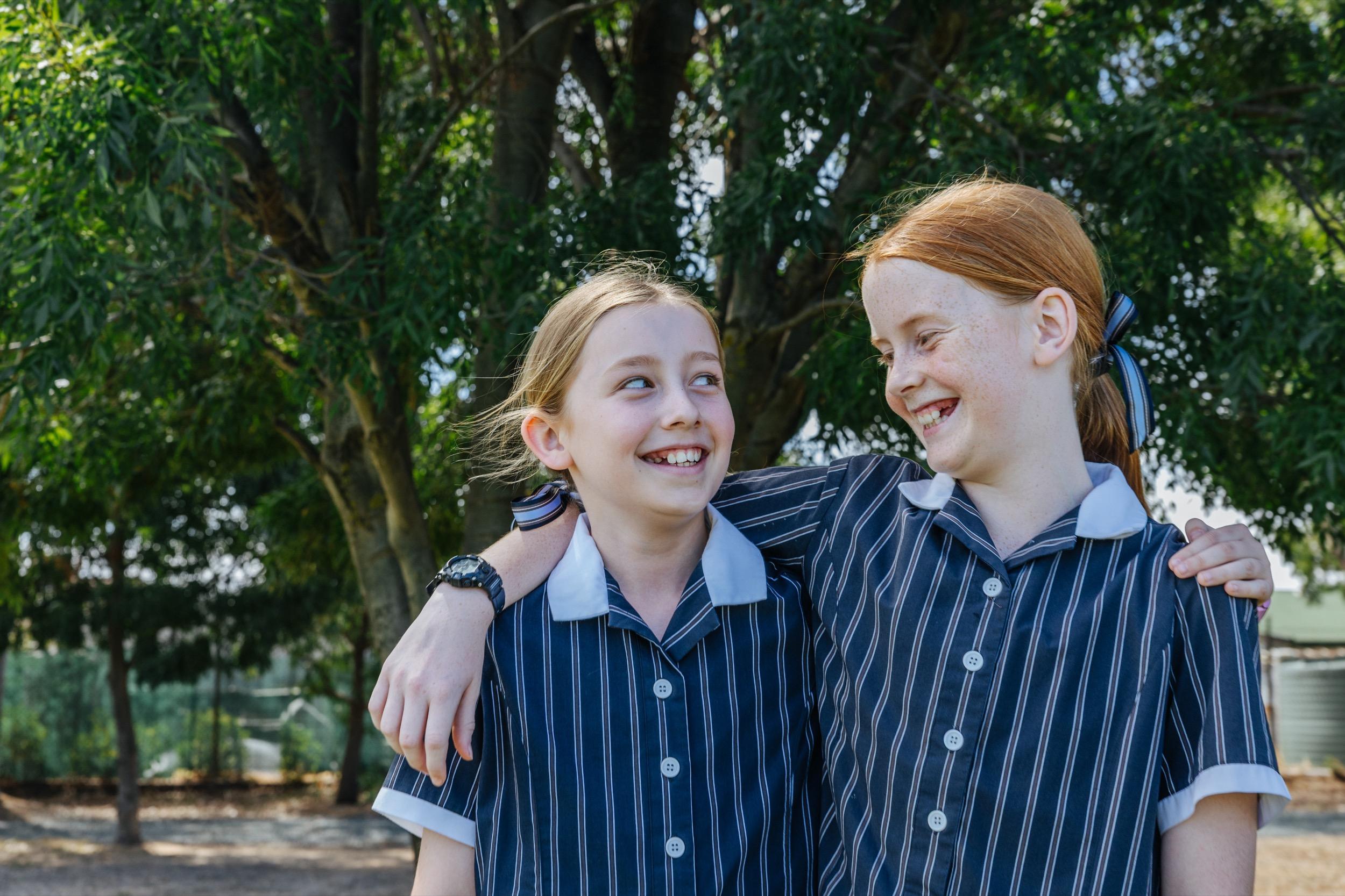 Two students in school uniforms stand outdoors, smiling and looking at each other while embracing each other’s shoulders.