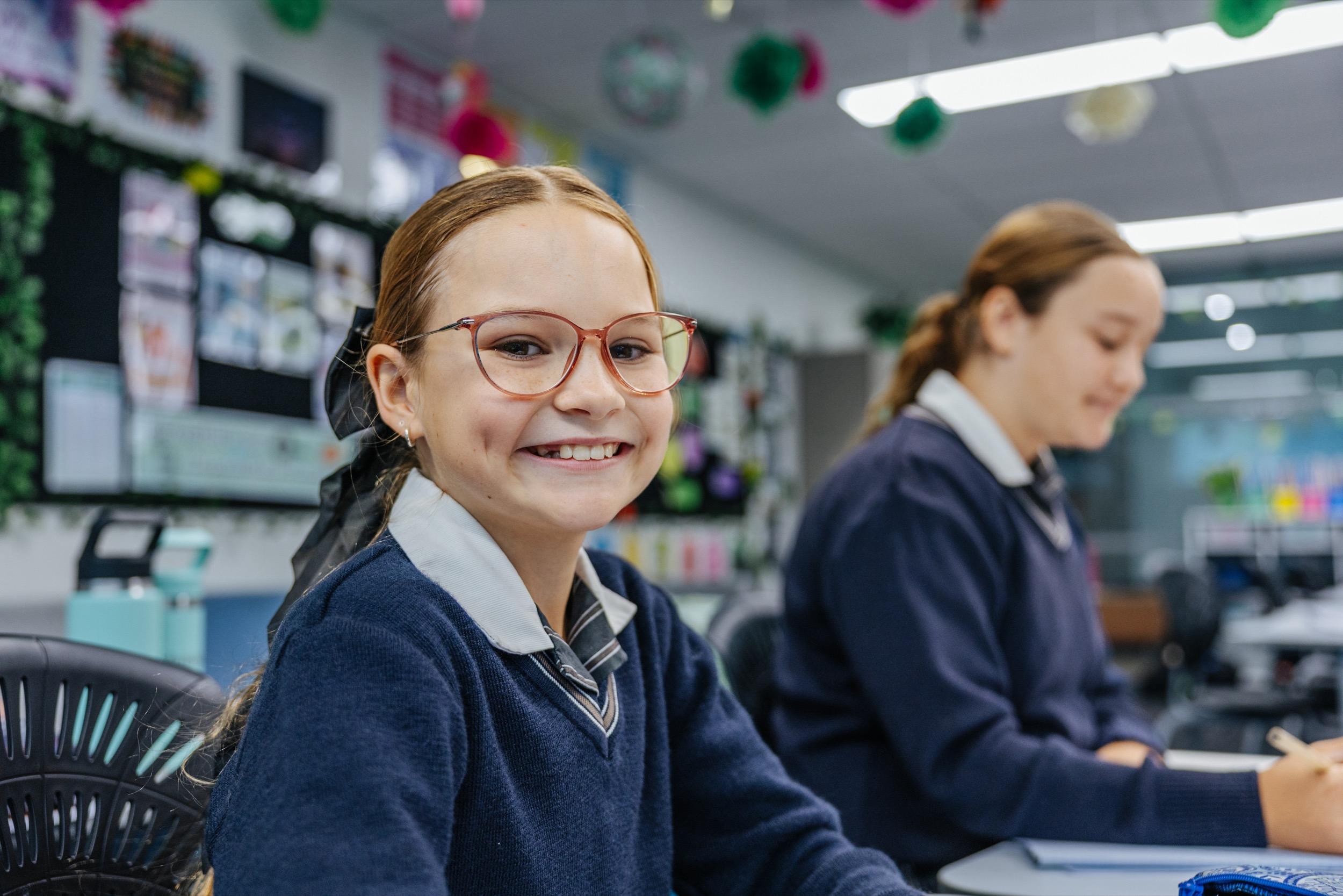 A student with glasses smiles at the camera while another student works at a desk in a classroom.