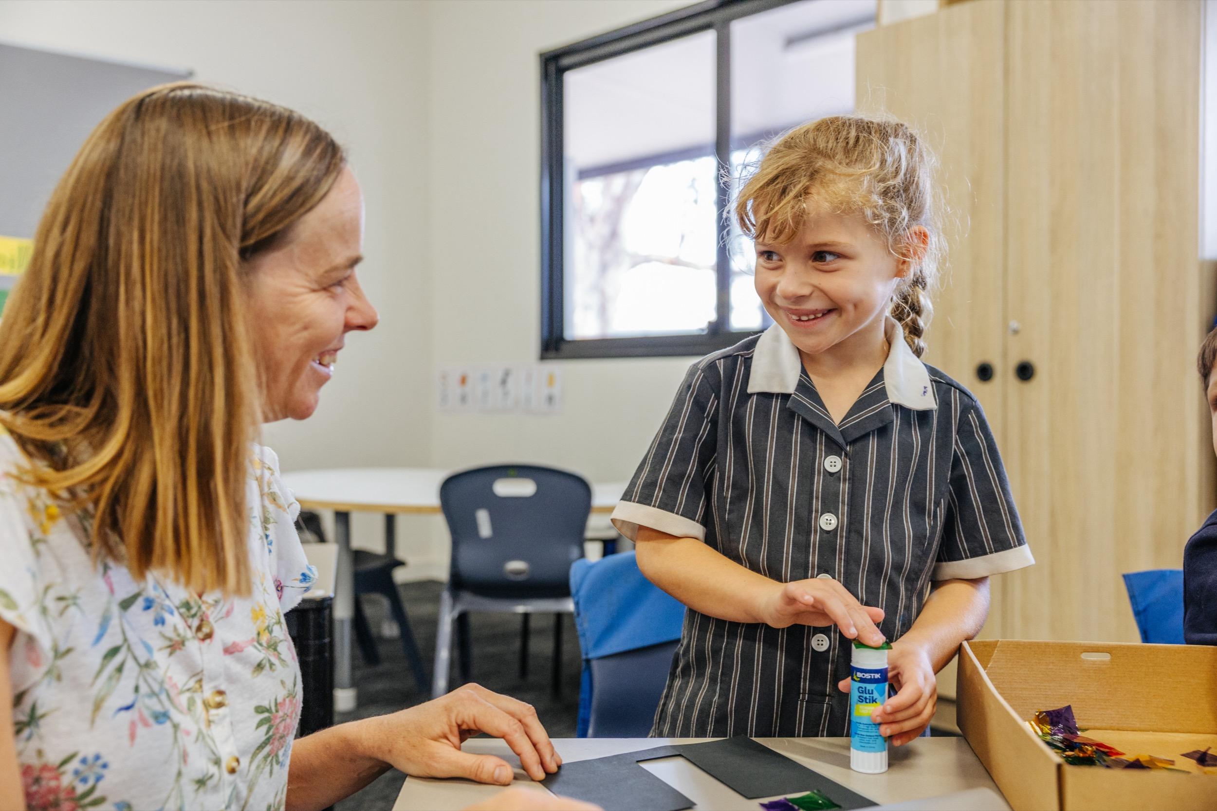 A teacher and a student smile at each other while working on a craft project at a classroom