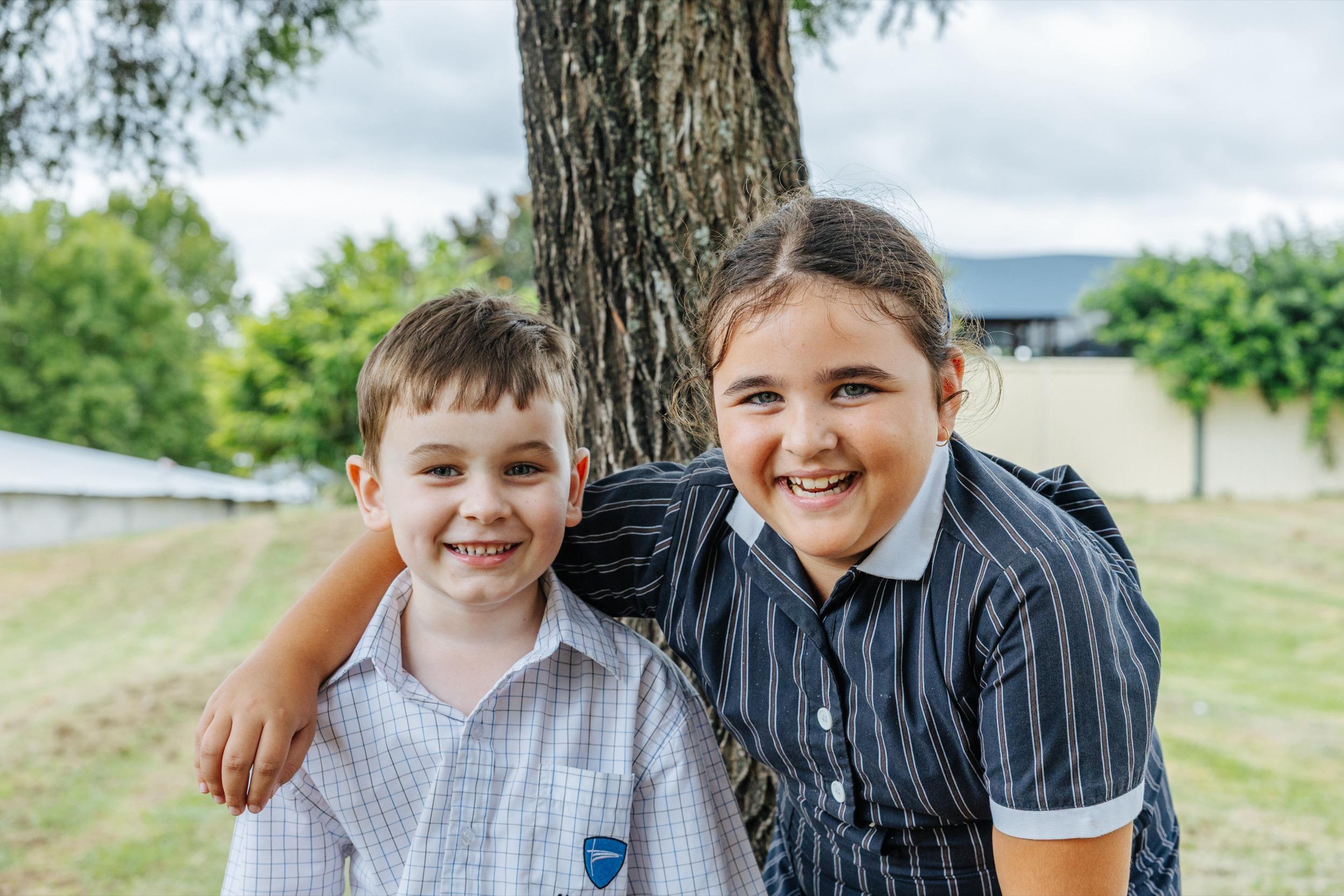Two students, a boy and a girl, smile while standing together by a tree in a grassy area.