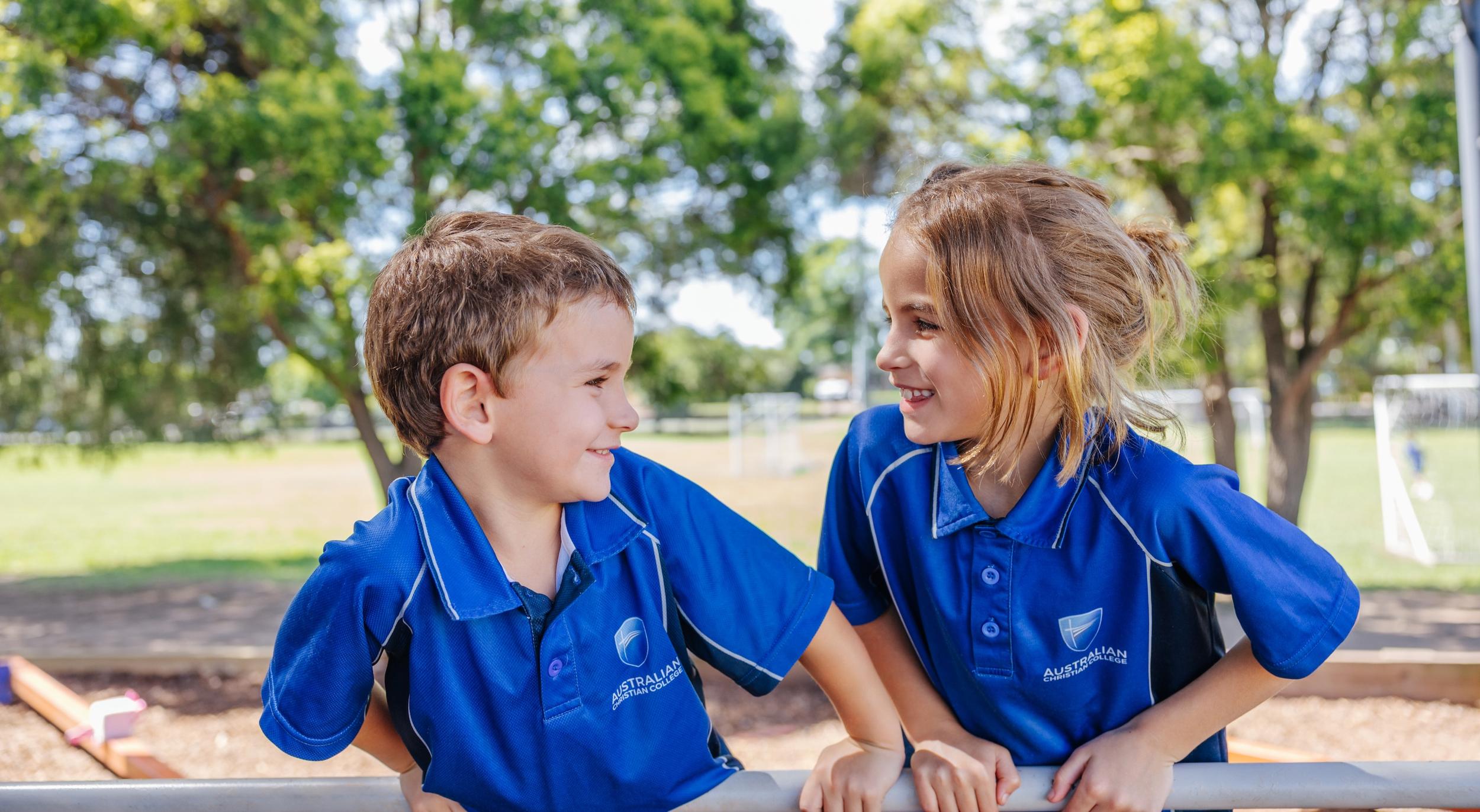 Two students in school uniforms smile at each other while leaning on a railing in a playground