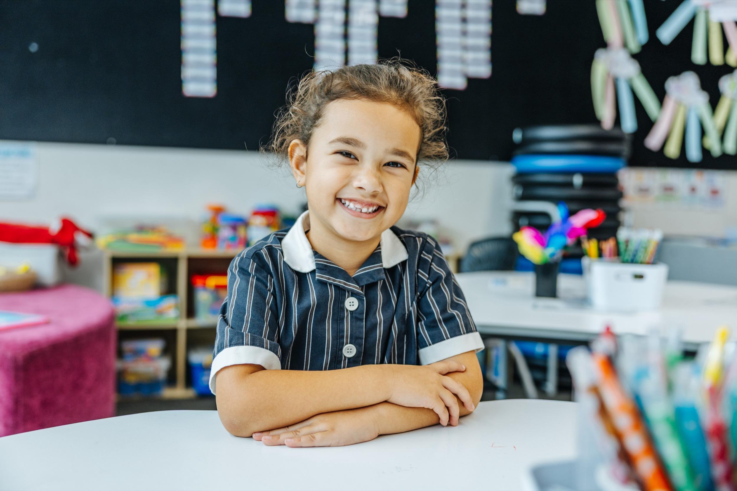 A smiling student in a school uniform sits at a table, with colorful stationery and classroom materials