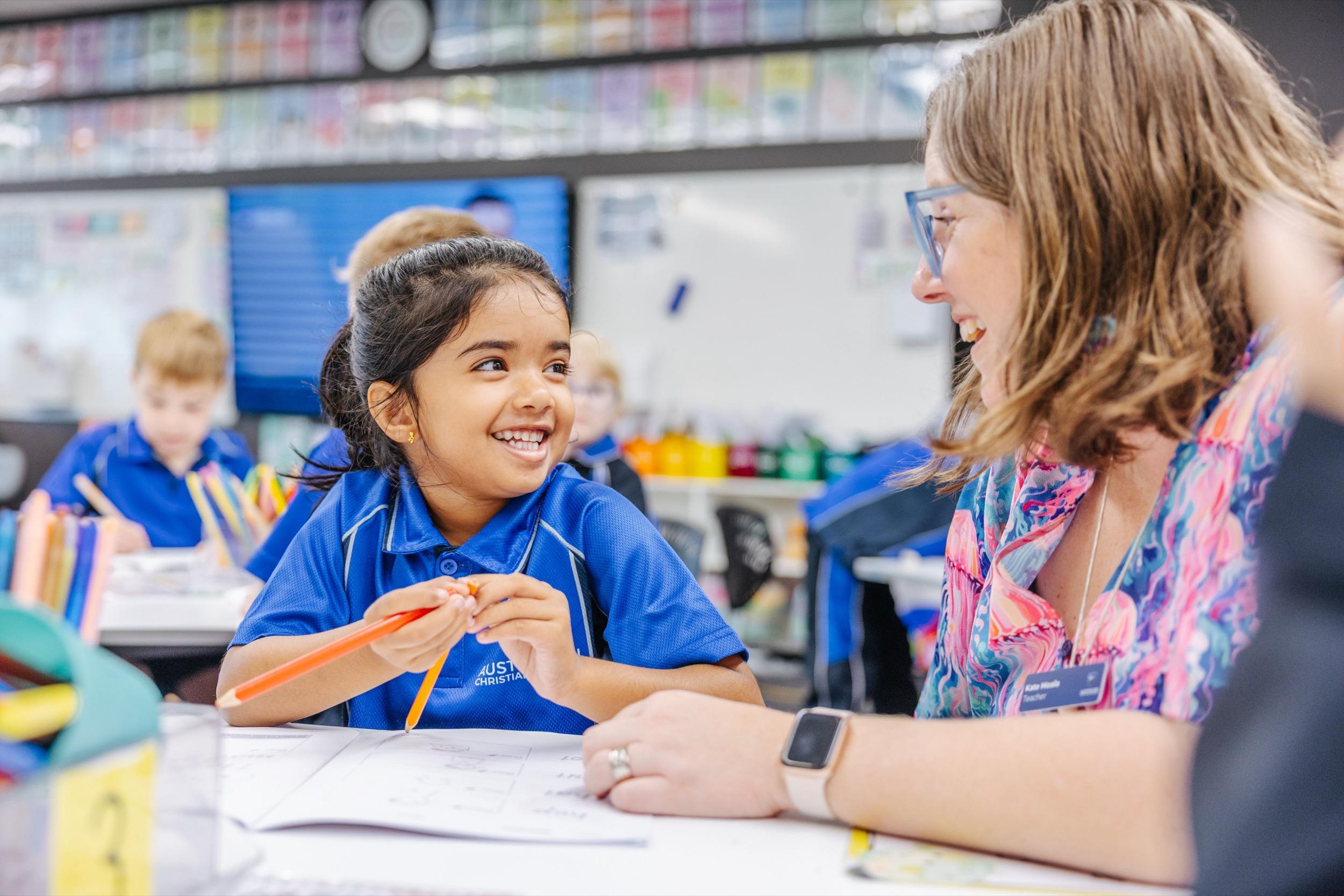 A student smiles while interacting with a teacher in a classroom, surrounded by colorful learning materials and other