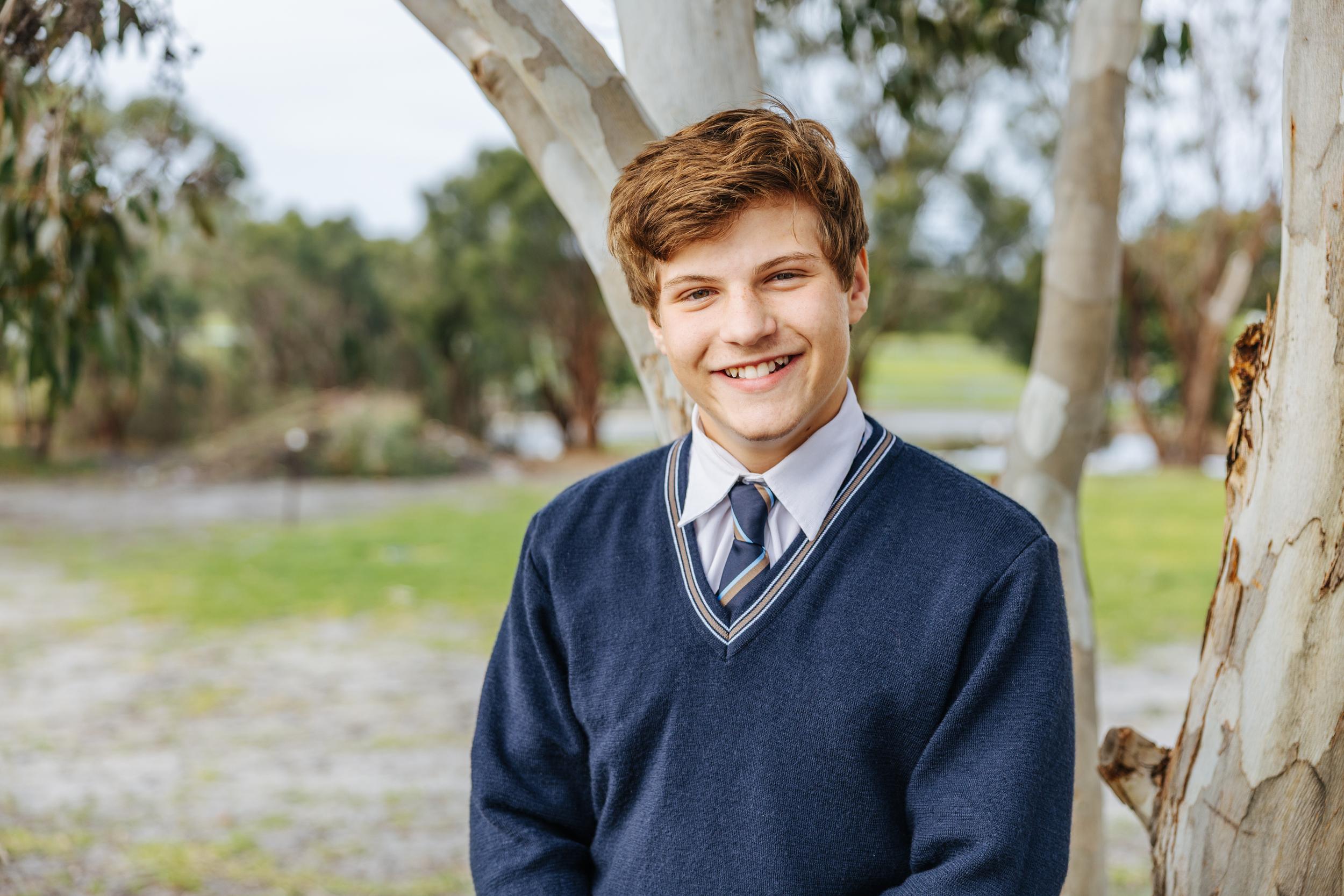 Student in school uniform smiling while standing next to a tree in a grassy outdoor setting.