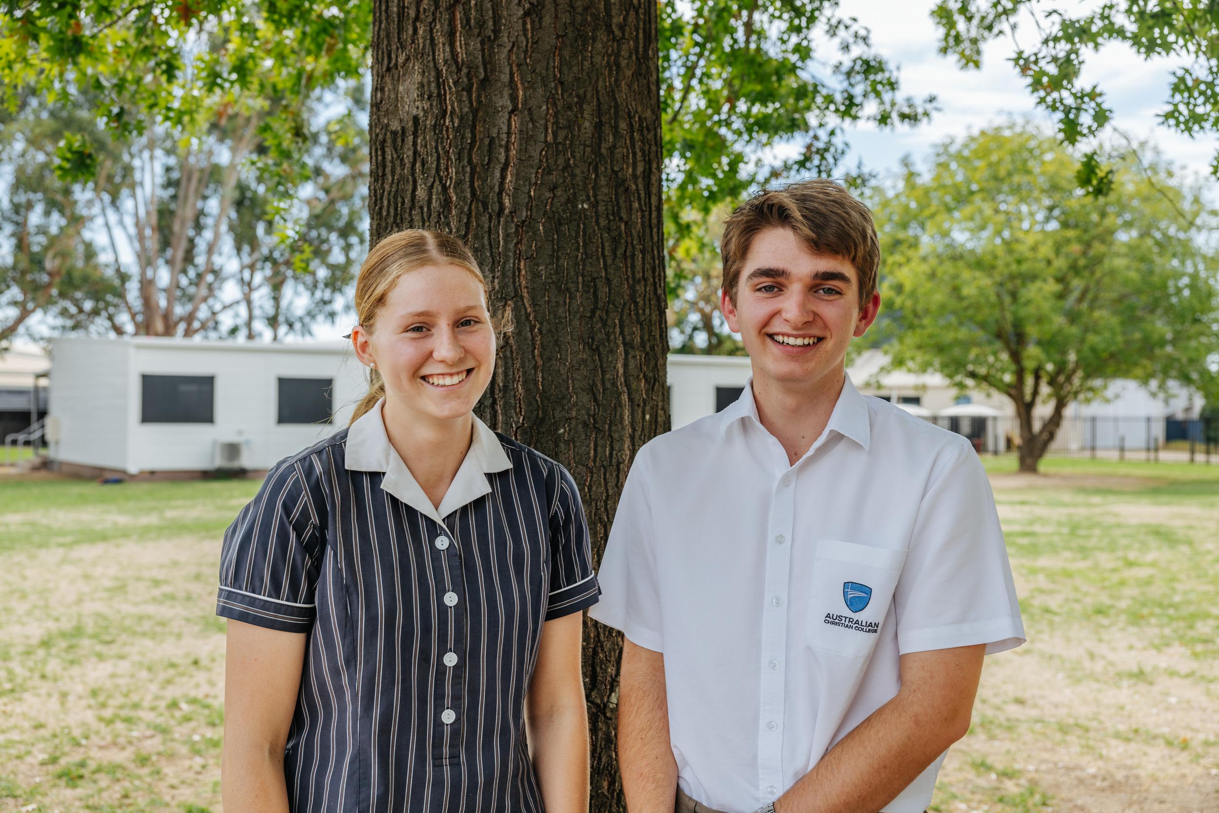 Students in school uniforms stand beside a tree, smiling at the camera, with classrooms visible in the background