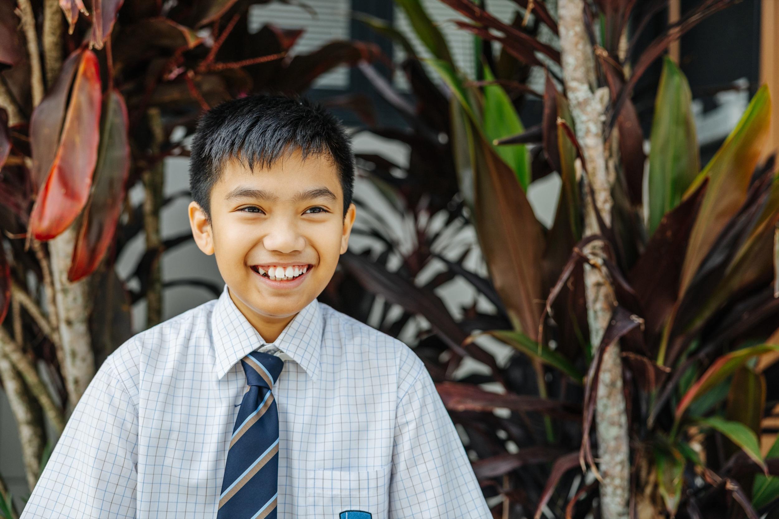Smiling student in school uniform stands in front of large green and red plants, expressing happiness.