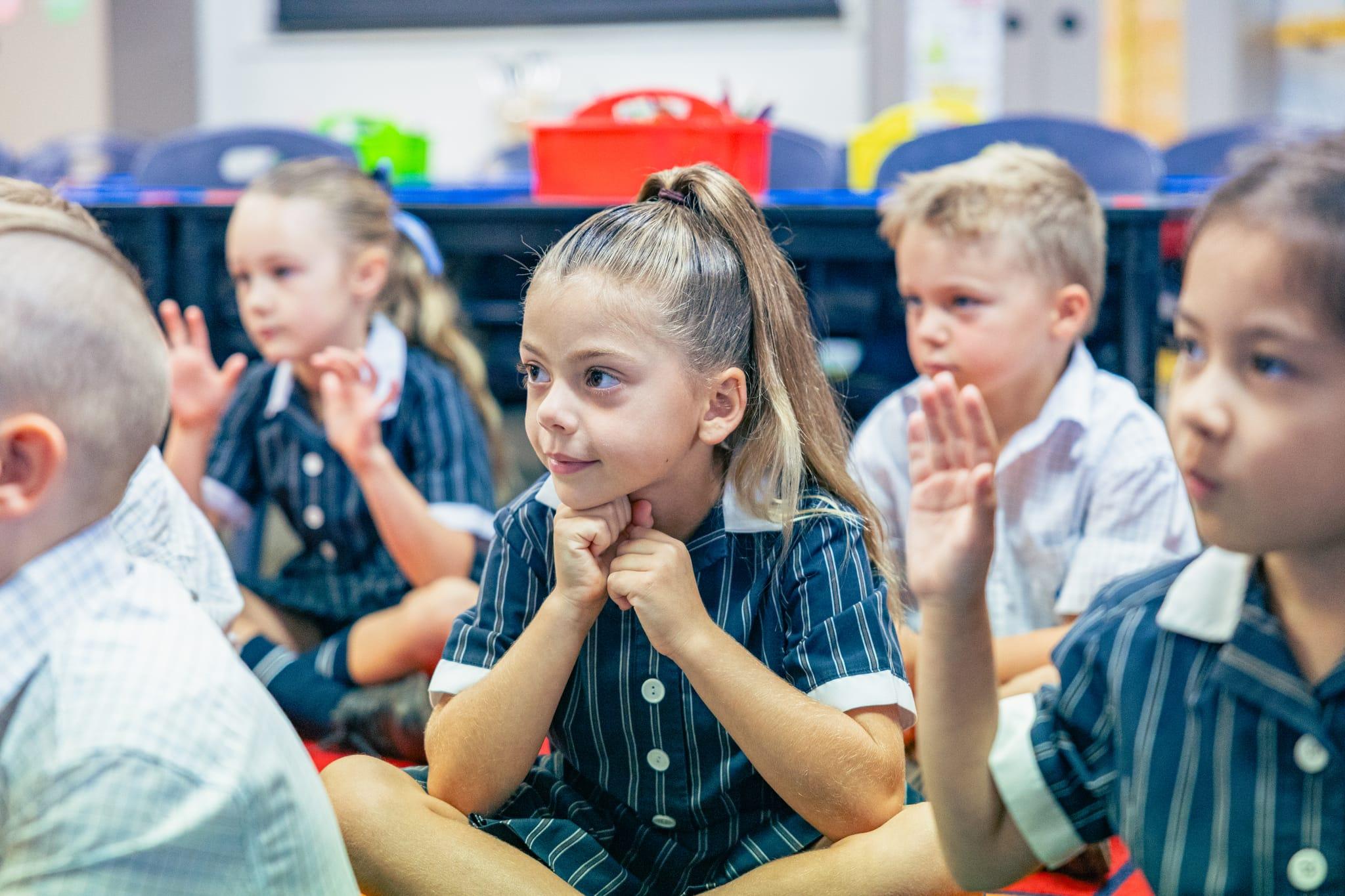 Students sitting on the floor in a classroom, engaged and raising their hands while listening attentively.