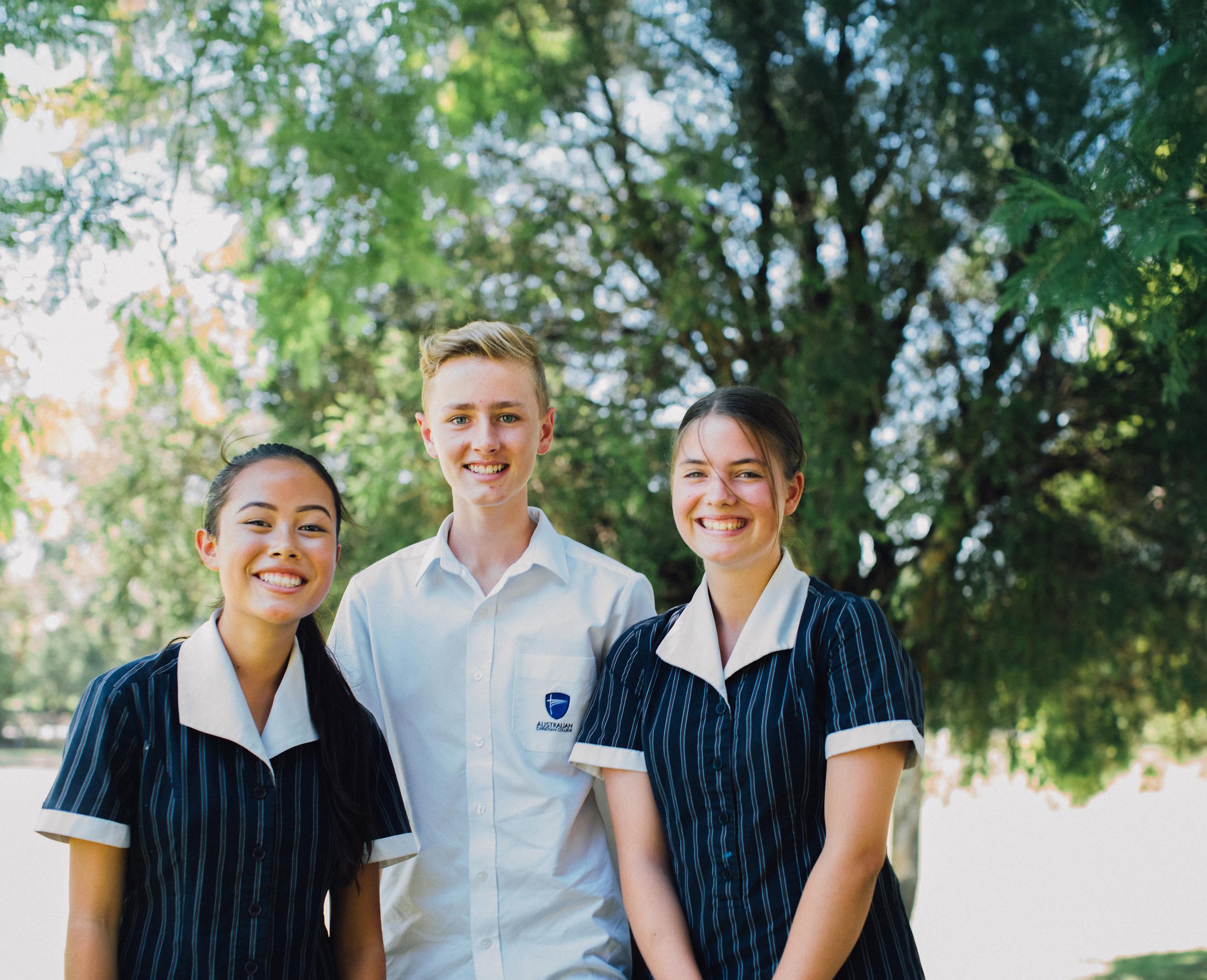 Students in school uniforms stand beside a tree, smiling at the camera, with classrooms visible in the background