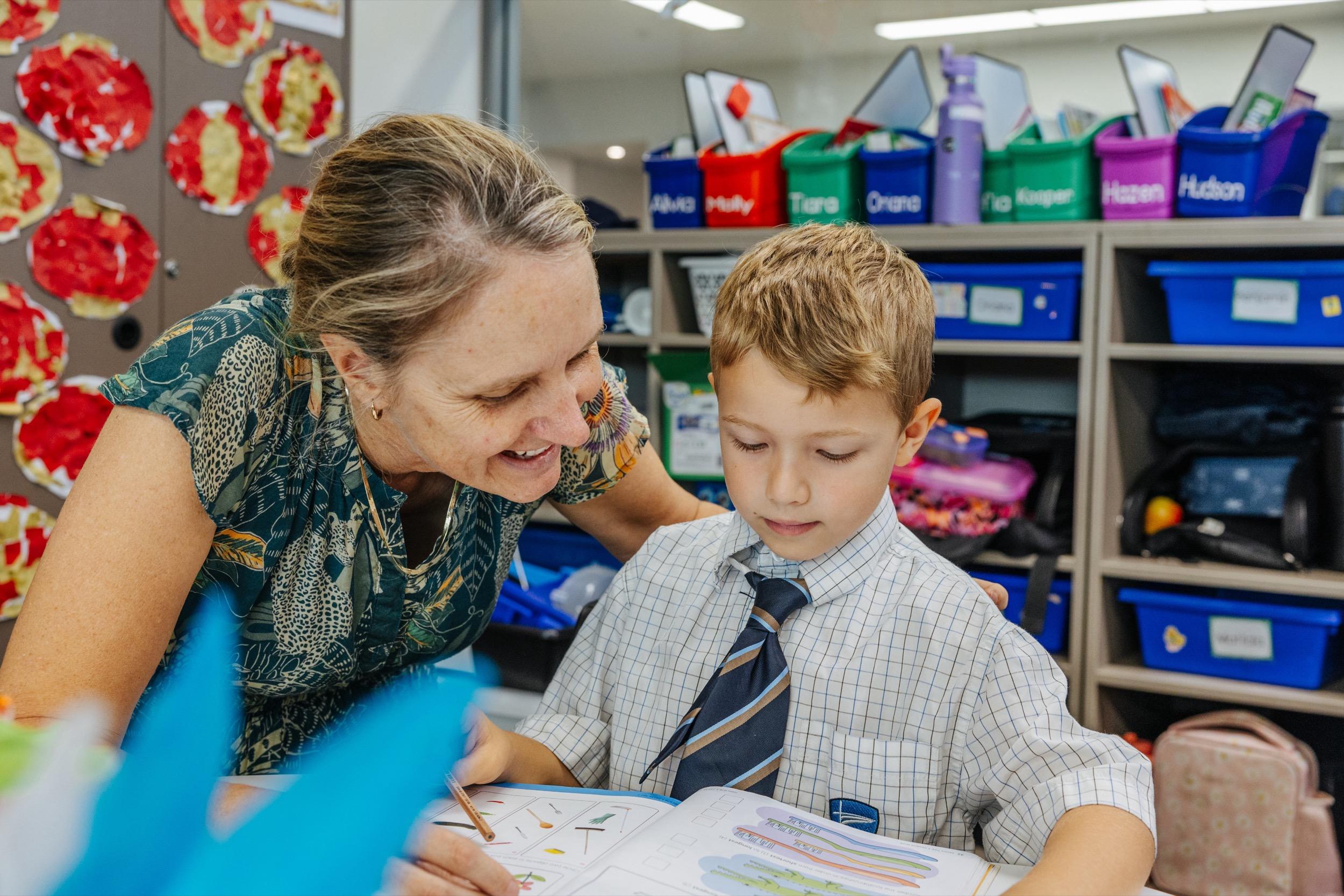 A teacher assists a young student with his work at a classroom desk, surrounded by colorful storage bins.