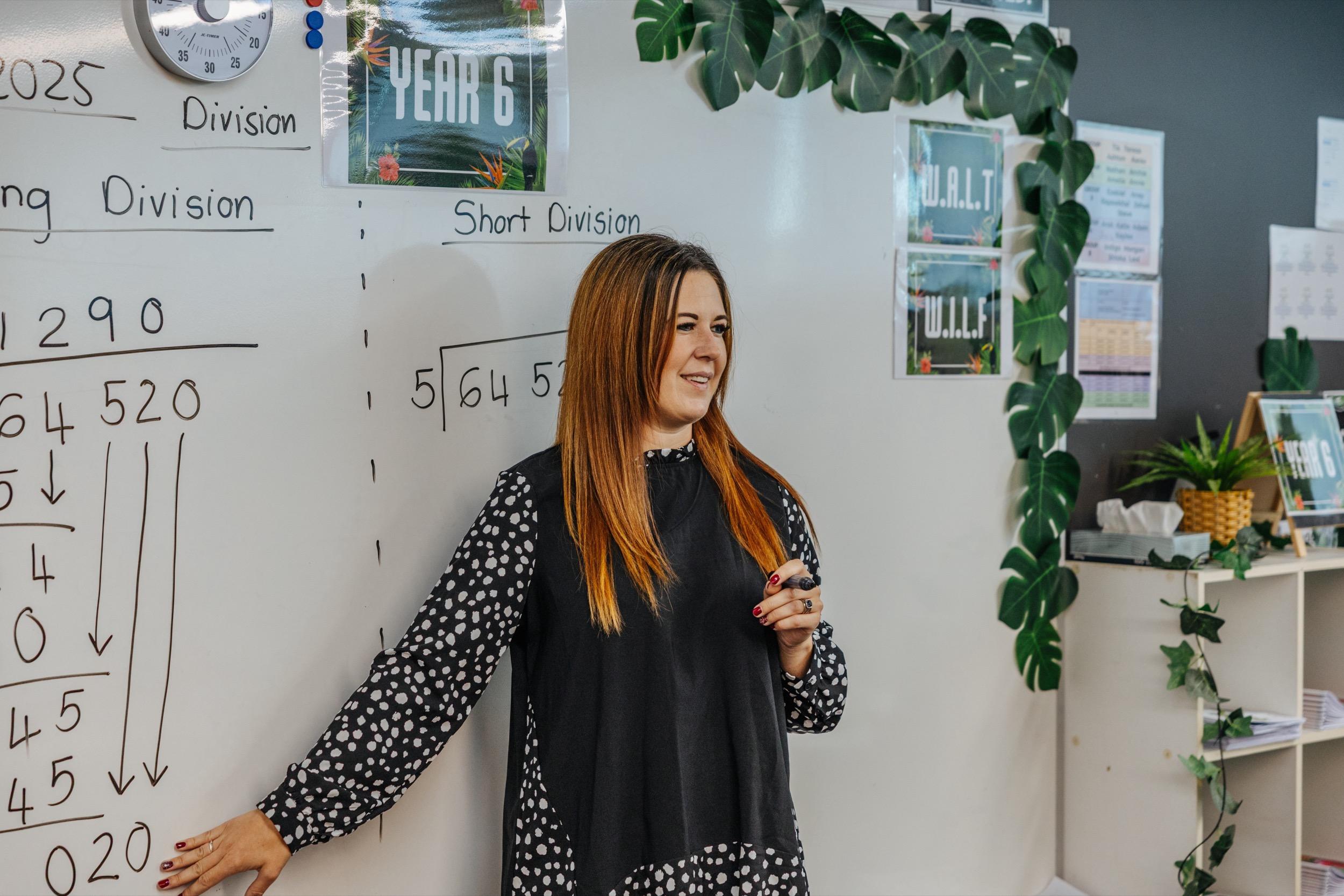 Teacher explaining division concepts at a whiteboard, with math problems written out and educational posters in the background.
