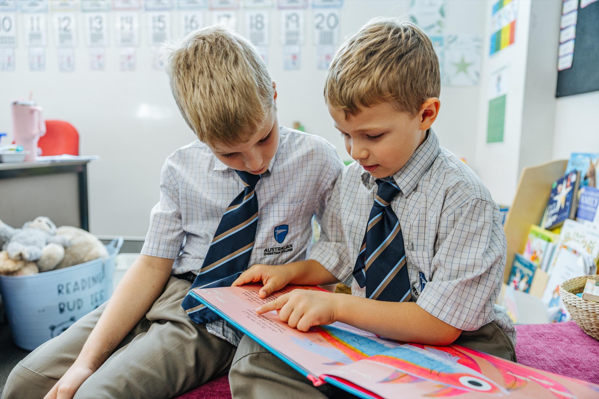 Two students in school uniform sit closely together, reading a book and pointing at the pages.