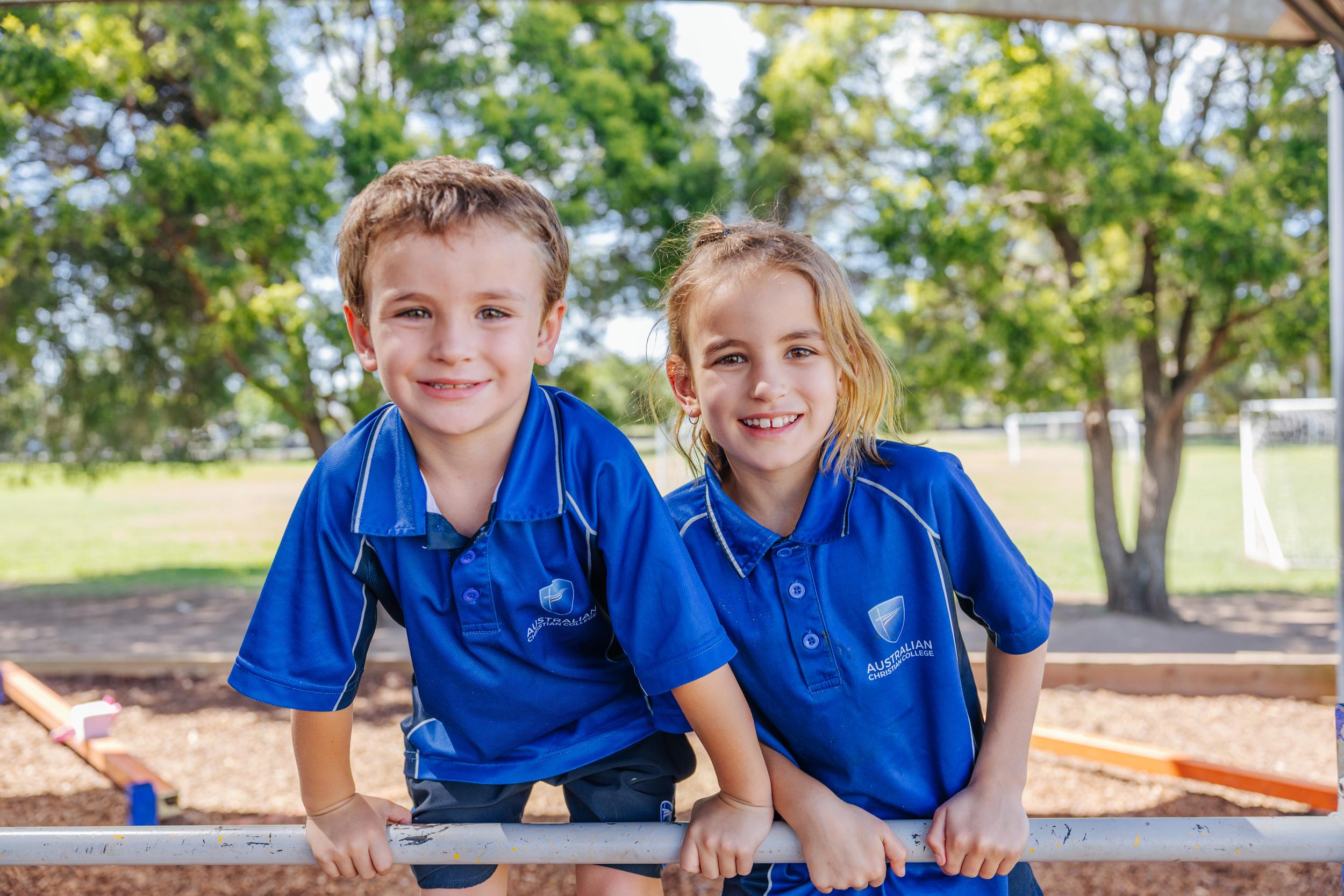 Two students in school uniform smile while sitting on a playground bar, with trees and a sports field