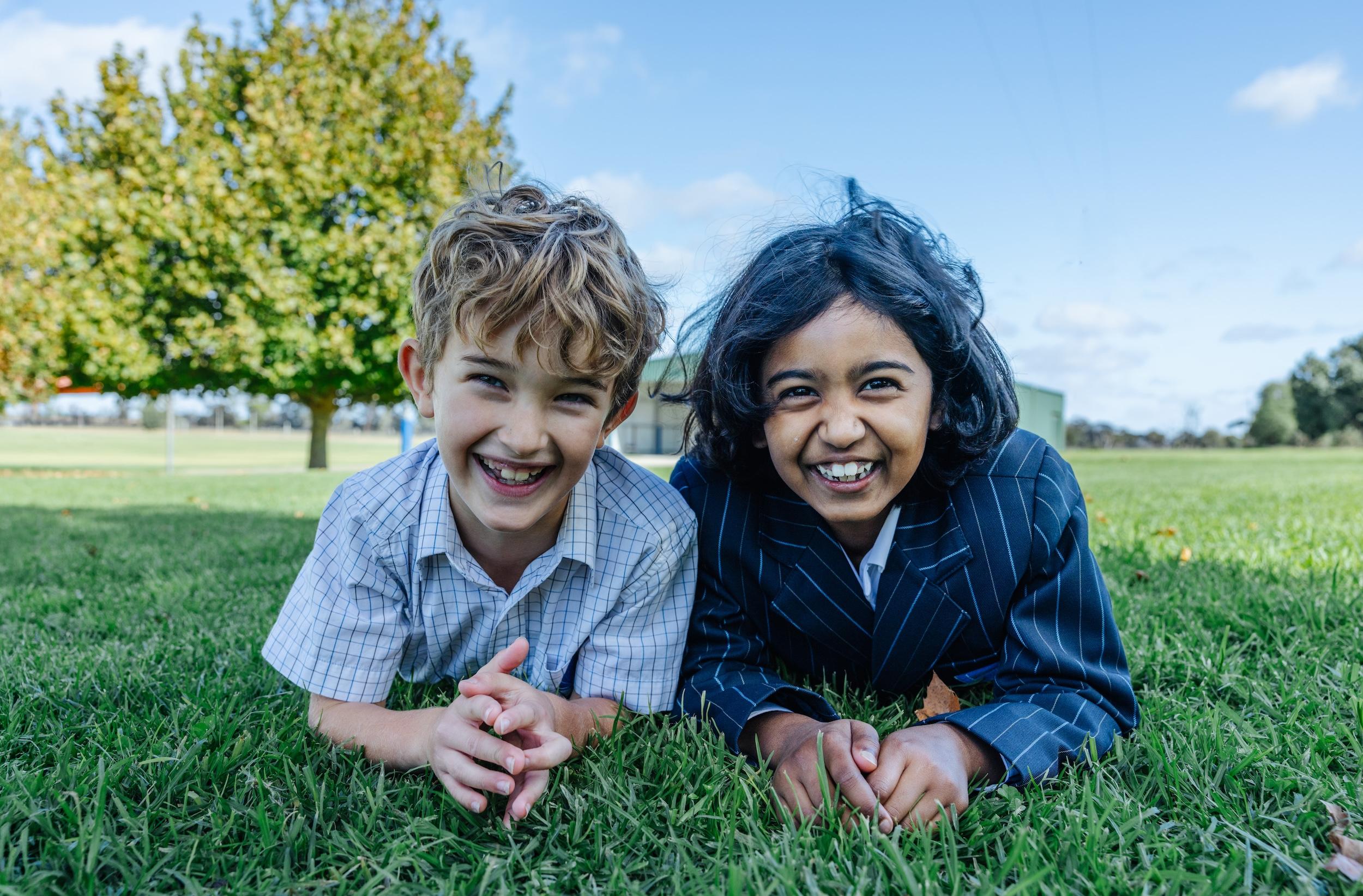 Two girls in school uniforms lying on grass, smiling at the camera, with trees in the background