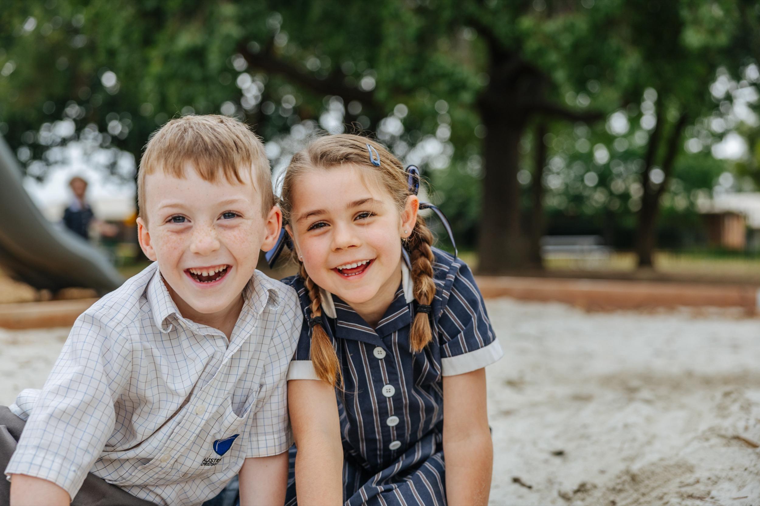 Two students smiling and sitting together in a sandpit, wearing school uniforms. Trees and playground equipment are visible